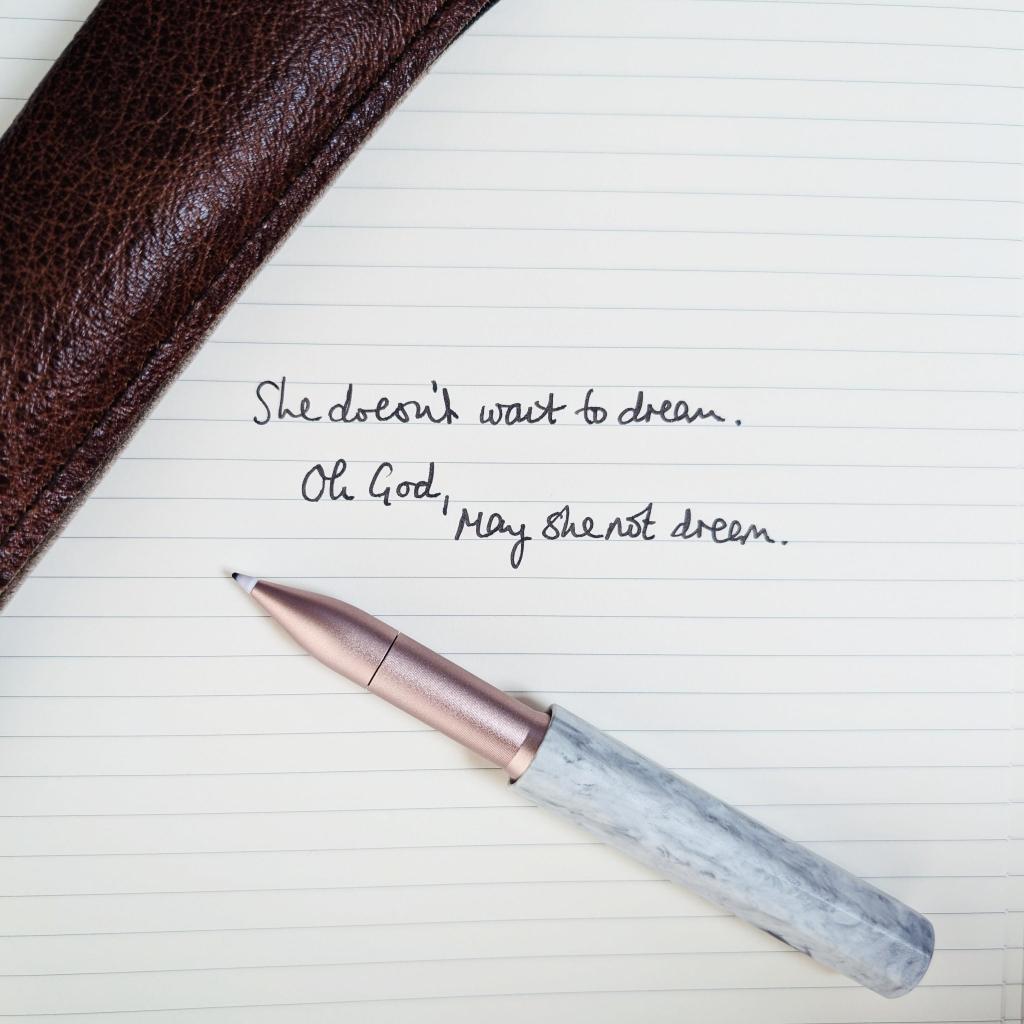 A close-up of an open notebook with handwritten text reading 'She doesn’t want to dream. Oh God, may she not dream.' alongside a stylish pen resting on the page and a brown leather case in the background.
